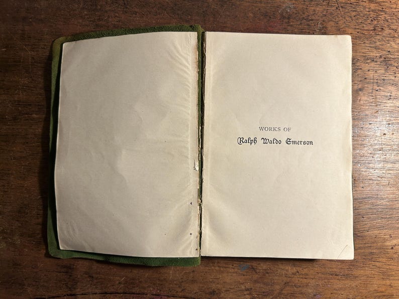 Pode incluir: Livro aberto com o t&iacute;tulo "WORKS OF Ralph Waldo Emerson" impresso em tinta preta. As p&aacute;ginas s&atilde;o brancas sujas e a capa &eacute; verde escuro. O livro est&aacute; sobre uma superf&iacute;cie de madeira.