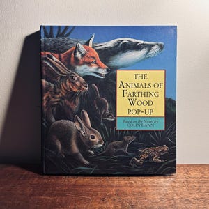 K&ouml;nnte beinhalten: Ein Pop-up-Buch mit dem Titel "The Animals of Farthing Wood" mit Illustrationen von Waldtieren. Das Cover zeigt einen Fuchs, einen Dachs, Kaninchen und einen Frosch vor blauem Himmel. Basierend auf dem Roman von Colin Dann.