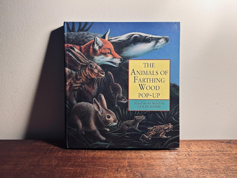 K&ouml;nnte beinhalten: Ein Pop-up-Buch mit dem Titel "The Animals of Farthing Wood" mit Illustrationen von Waldtieren. Das Cover zeigt einen Fuchs, einen Dachs, Kaninchen und einen Frosch vor blauem Himmel. Basierend auf dem Roman von Colin Dann.
