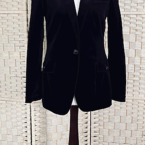 May include: A black velvet blazer displayed on a mannequin. The blazer has a notched lapel, two front pockets, and a single-button closure. The mannequin is on a dark wooden stand, set against a neutral background.