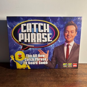 Puede incluir: Una caja del juego de mesa de televisión Catch Phrase. La caja es azul con el título del juego en letras blancas grandes. Un personaje de dibujos animados y un hombre con traje también están en la caja. El texto dice "The All New Catch Phrase TV Board Game."