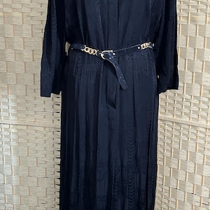 Puede incluir: Vestido largo negro con un patrón sutil, abotonado en el cuello y ceñido en la cintura con un cinturón negro con detalles de cadena dorada. El vestido se exhibe en un maniquí.