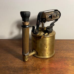 Pode incluir: Um ma&ccedil;arico de lat&atilde;o vintage com al&ccedil;a e bico pretos. O ma&ccedil;arico tem um tanque de metal e uma correia de couro preta.