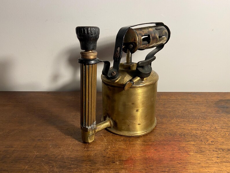 Pode incluir: Um ma&ccedil;arico de lat&atilde;o vintage com al&ccedil;a e bico pretos. O ma&ccedil;arico tem um tanque de metal e uma correia de couro preta.