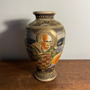 Può includere: Un piccolo vaso in ceramica decorativo con un design oro e nero. Il vaso presenta un'immagine dipinta di un uomo con la barba e una corona. Il vaso è alto circa 15 cm.