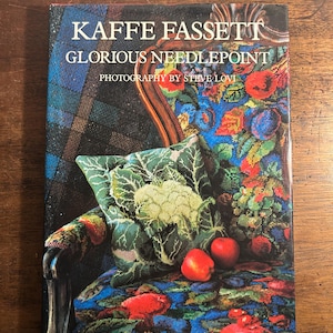 May include: The book cover of "KAFFE FASSETT GLORIOUS NEEDLEPOINT" with photography by Steve Lovi. It shows a vibrant needlepoint design of a chair, a cushion with a cauliflower motif, and two red apples.