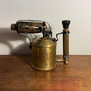 Pode incluir: Um ma&ccedil;arico vintage de lat&atilde;o com uma pega preta e um bico de metal. O ma&ccedil;arico tem um r&oacute;tulo com instru&ccedil;&otilde;es de uso.