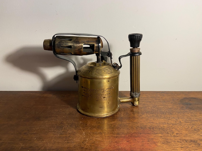 Pode incluir: Um ma&ccedil;arico vintage de lat&atilde;o com uma pega preta e um bico de metal. O ma&ccedil;arico tem um r&oacute;tulo com instru&ccedil;&otilde;es de uso.