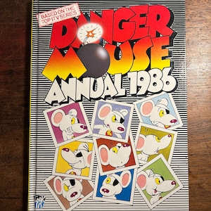 Op de afbeelding: Een vintage "Danger Mouse Annual 1986" boek. De cover toont de titel in rood en geel, een bomafbeelding en afbeeldingen van de cartoonmuis. Het boek is gebaseerd op de Top ITV-serie.