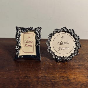 Peut inclure: Deux petits cadres photo décoratifs. L'un est rectangulaire avec des ornements floraux et de feuilles, l'autre est rond avec une bordure en cristal. Les deux cadres ont une insertion crème avec le texte "A Classic Frame".