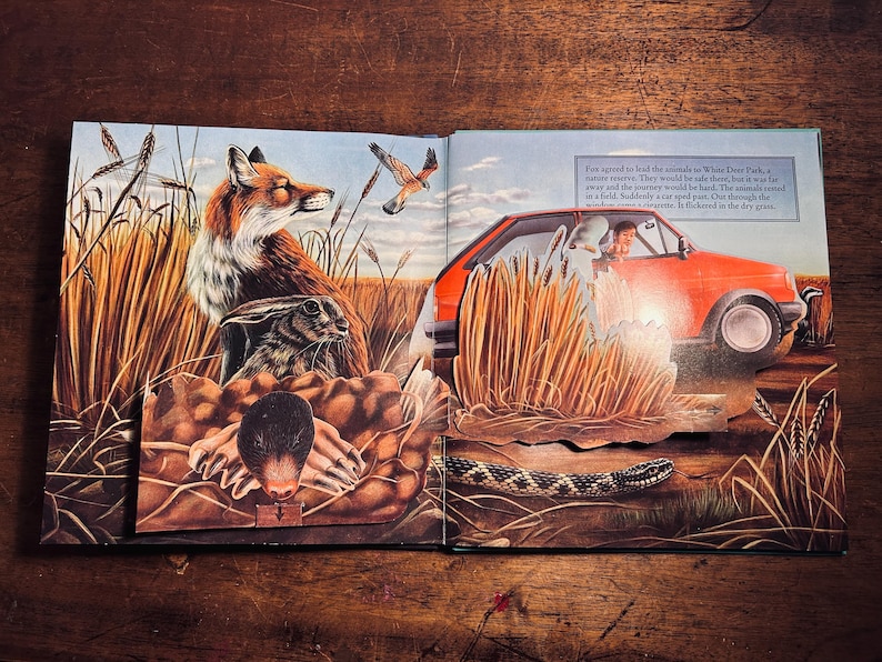 K&ouml;nnte beinhalten: Illustration eines offenen Buches mit einem Fuchs, einem Kaninchen und einem Vogel auf einem Feld. Rechts ist ein rotes Auto mit einer Person darin zu sehen. Der Text lautet: "Fox stimmte zu, die Tiere zum White Deer Park zu f&uuml;hren..." und "Pl&ouml;tzlich raste ein Auto vorbei."