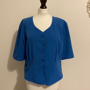 Op de afbeelding: Een blauwe blouse met korte mouwen, een V-hals en een knoopsluiting, gepresenteerd op een mannequin. De mouwen zijn licht gecapt en de taille is zacht getailleerd. De stof heeft een gladde, effen uitstraling, kenmerkend voor een klassiek ontwerp.