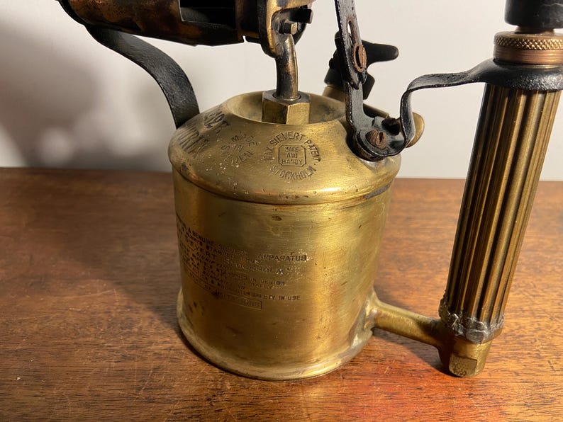 Pode incluir: Um ma&ccedil;arico vintage de lat&atilde;o com cabo preto e bico de lat&atilde;o. O ma&ccedil;arico tem um r&oacute;tulo que diz "S.L.Sivert Patent" e "Estocolmo".