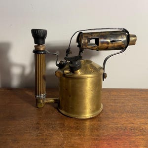 Pode incluir: Um ma&ccedil;arico de lat&atilde;o vintage com uma pega preta e um bico de metal. O ma&ccedil;arico est&aacute; sobre uma superf&iacute;cie de madeira.