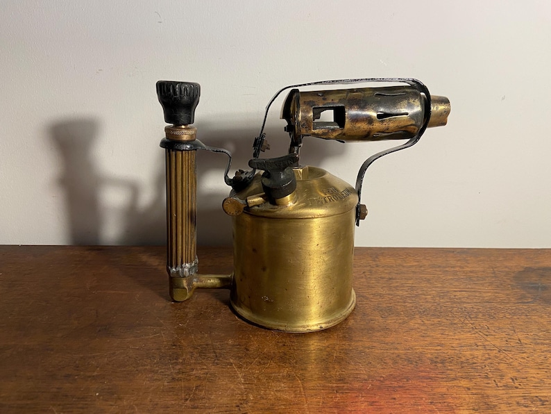 Pode incluir: Um ma&ccedil;arico de lat&atilde;o vintage com uma pega preta e um bico de metal. O ma&ccedil;arico est&aacute; sobre uma superf&iacute;cie de madeira.