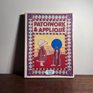 May include: A vintage book titled "PATCHWORK & APPLIQUE" by Pamela Tubby. The cover features a colourful illustration of two figures in a field, with a patchwork border in red, white, and blue. The book is on a wooden surface.