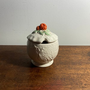 Puede incluir: Un azucarero de cerámica blanca con tapa, con un diseño de hoja de col. La tapa está rematada con un racimo de bayas naranjas y hojas verdes. El cuenco está sobre una superficie de madera.
