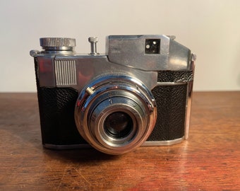 Puede incluir: Una cámara de película vintage de 35 mm de color plateado y negro con una lente marcada con "70" y una cubierta de cuero negro.