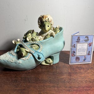 May include: A light blue ceramic shoe figurine containing several small mice, with one larger mouse wearing a bonnet. The shoe features white lace-like detailing. Beside it stands a small booklet titled The World of Beatrix Potter.