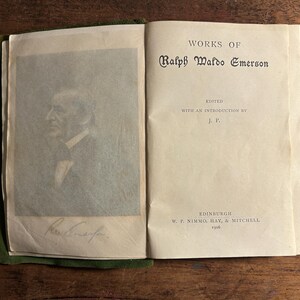 Pode incluir: Livro aberto com o t&iacute;tulo "WORKS OF Ralph Waldo Emerson" impresso na p&aacute;gina direita. A p&aacute;gina esquerda apresenta um retrato e uma assinatura. O livro est&aacute; encadernado em verde e repousa sobre uma superf&iacute;cie de madeira.