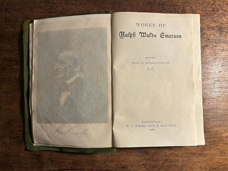 Pode incluir: Livro aberto com o t&iacute;tulo "WORKS OF Ralph Waldo Emerson" impresso na p&aacute;gina direita. A p&aacute;gina esquerda apresenta um retrato e uma assinatura. O livro est&aacute; encadernado em verde e repousa sobre uma superf&iacute;cie de madeira.