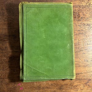 Pode incluir: Um livro antigo fechado com uma capa verde. A capa &eacute; de uma cor verde lisa e discreta com uma borda retangular. As bordas do livro est&atilde;o desgastadas, mostrando sinais de idade. O livro est&aacute; sobre uma superf&iacute;cie de madeira.