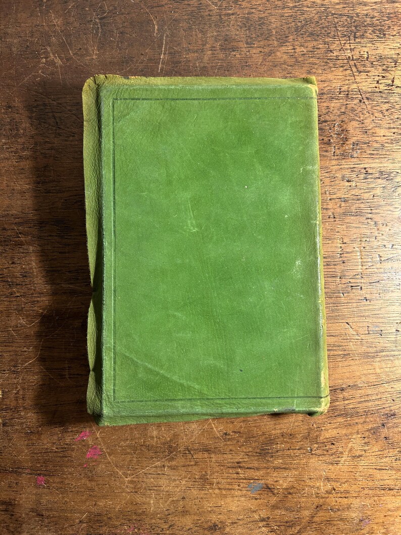 Pode incluir: Um livro antigo fechado com uma capa verde. A capa &eacute; de uma cor verde lisa e discreta com uma borda retangular. As bordas do livro est&atilde;o desgastadas, mostrando sinais de idade. O livro est&aacute; sobre uma superf&iacute;cie de madeira.