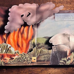 K&ouml;nnte beinhalten: Illustration eines Pop-up-Buchs, das einen Waldbrand mit einem laufenden Fuchs zeigt. Die Szene umfasst hohes Gras, ein gro&szlig;es orangefarbenes Feuer und eine graue Wolke. Der Text auf der rechten Seite lautet: "Fuchs' Nase zuckte vor Schreck."