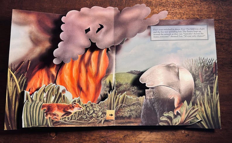 K&ouml;nnte beinhalten: Illustration eines Pop-up-Buchs, das einen Waldbrand mit einem laufenden Fuchs zeigt. Die Szene umfasst hohes Gras, ein gro&szlig;es orangefarbenes Feuer und eine graue Wolke. Der Text auf der rechten Seite lautet: "Fuchs' Nase zuckte vor Schreck."