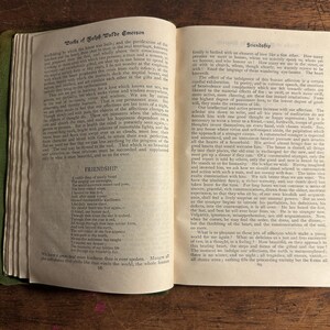Pode incluir: Um livro aberto com bordas verdes, exibindo texto de "Works of Ralph Waldo Emerson" e o poema "Friendship". As p&aacute;ginas est&atilde;o envelhecidas, com texto vis&iacute;vel e uma est&eacute;tica vintage. O livro est&aacute; em uma superf&iacute;cie de madeira.