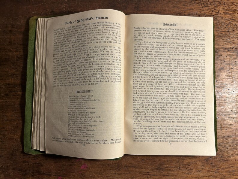 Pode incluir: Um livro aberto com bordas verdes, exibindo texto de "Works of Ralph Waldo Emerson" e o poema "Friendship". As p&aacute;ginas est&atilde;o envelhecidas, com texto vis&iacute;vel e uma est&eacute;tica vintage. O livro est&aacute; em uma superf&iacute;cie de madeira.