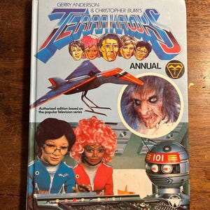 Puede incluir: Una portada de libro vintage "Terra Hawks Annual". La portada presenta ilustraciones de personajes, una aeronave futurista y un rostro amenazante. El libro es una edición autorizada basada en la popular serie de televisión.