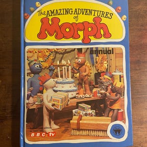 Op de afbeelding: Een kleurrijke boekomslag met de titel "The Amazing Adventures of Morph annual". De omslag toont een groep klei-animatiefiguren die een verjaardag vieren met een taart en cadeaus. Het boek is een geautoriseerde editie zoals te zien op BBC TV.