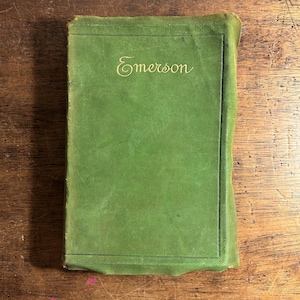 Pode incluir: Um livro verde vintage com a palavra "Emerson" em dourado na capa. O livro tem formato retangular e est&aacute; sobre uma superf&iacute;cie de madeira. A capa mostra sinais de desgaste.