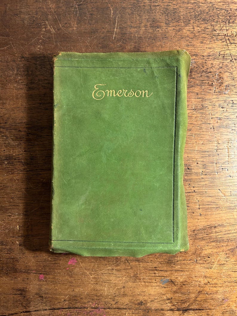 Pode incluir: Um livro verde vintage com a palavra "Emerson" em dourado na capa. O livro tem formato retangular e est&aacute; sobre uma superf&iacute;cie de madeira. A capa mostra sinais de desgaste.