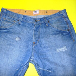 Puede incluir: Un par de pantalones vaqueros azules con una cintura marrón y una pequeña etiqueta que dice "Lee 101B" en la parte delantera. Los vaqueros tienen un aspecto desgastado con áreas descoloridas y algunas roturas.