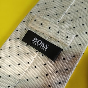 May include: A light beige necktie featuring a repeating pattern of small black squares. The tie includes a black fabric label stitched onto the back that displays the white text BOSS HUGO BOSS. The material has a visible woven texture.