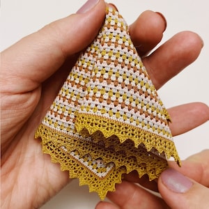 Könnte beinhalten: Gefaltetes gehäkeltes Textil in Gold-, Weiß-, Rosa- und Brauntönen. Die Kanten sind mit einem goldfarbenen, gewellten Rand versehen. Das offene Webmuster ist im gesamten Stoff sichtbar.