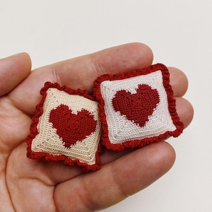 May include: Two miniature crochet pillows, each featuring a red heart on a cream and white background. The pillows have a ruffled red border. These handmade decorative items are perfect for dollhouses or craft projects.