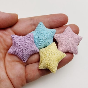 May include: Five crocheted star-shaped ornaments in pastel colors: lavender, light blue, yellow, and pink. Each star has a detailed openwork design. The stars are held in a hand, showcasing their size and texture.