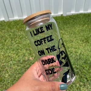 May include: A clear glass coffee tumbler with a bamboo lid. The tumbler features the text "I LIKE MY COFFEE ON THE DARK SIDE" in black, along with a black graphic of a character. The tumbler is held against a background of green grass.