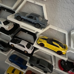 Può includere: Una collezione di macchinine giocattolo esposte su uno scaffale esagonale bianco. Le auto sono di vari colori, tra cui giallo, blu, rosso, bianco e grigio. Lo scaffale è montato su una parete grigia.