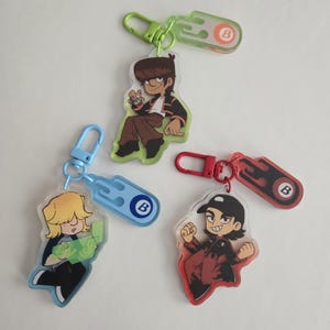 Puede incluir: Tres llaveros de personajes de dibujos animados con diseños coloridos. Cada llavero presenta un personaje diferente con una pose y un atuendo únicos. Los llaveros tienen una base de acrílico transparente y un cierre de metal. Los llaveros están unidos a una etiqueta de color a juego.