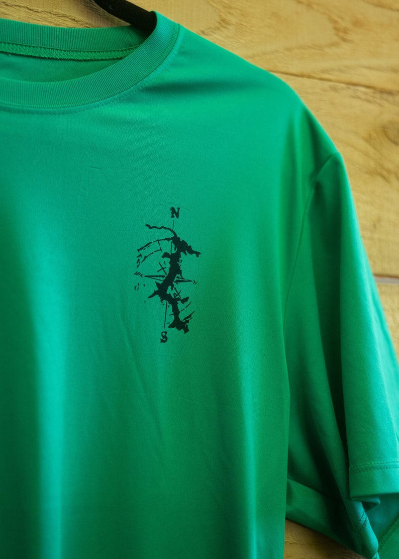 May include: A green t-shirt with a black compass design on the left side of the chest. The compass design features a stylised map with a north arrow and the letter "S" for south.