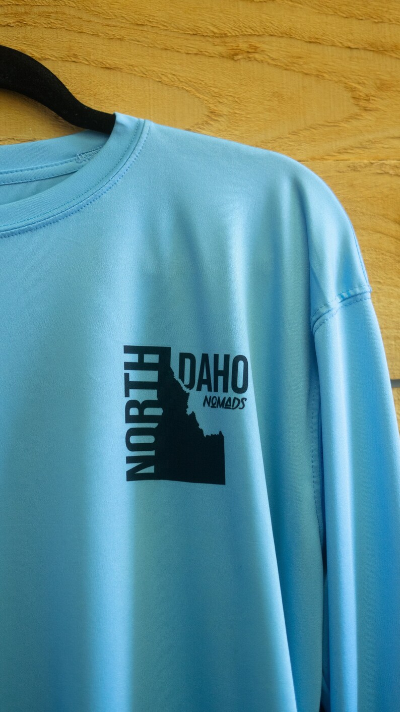 May include: A light blue long-sleeved shirt with a black graphic of the state of Idaho and the text "North Idaho Nomads".