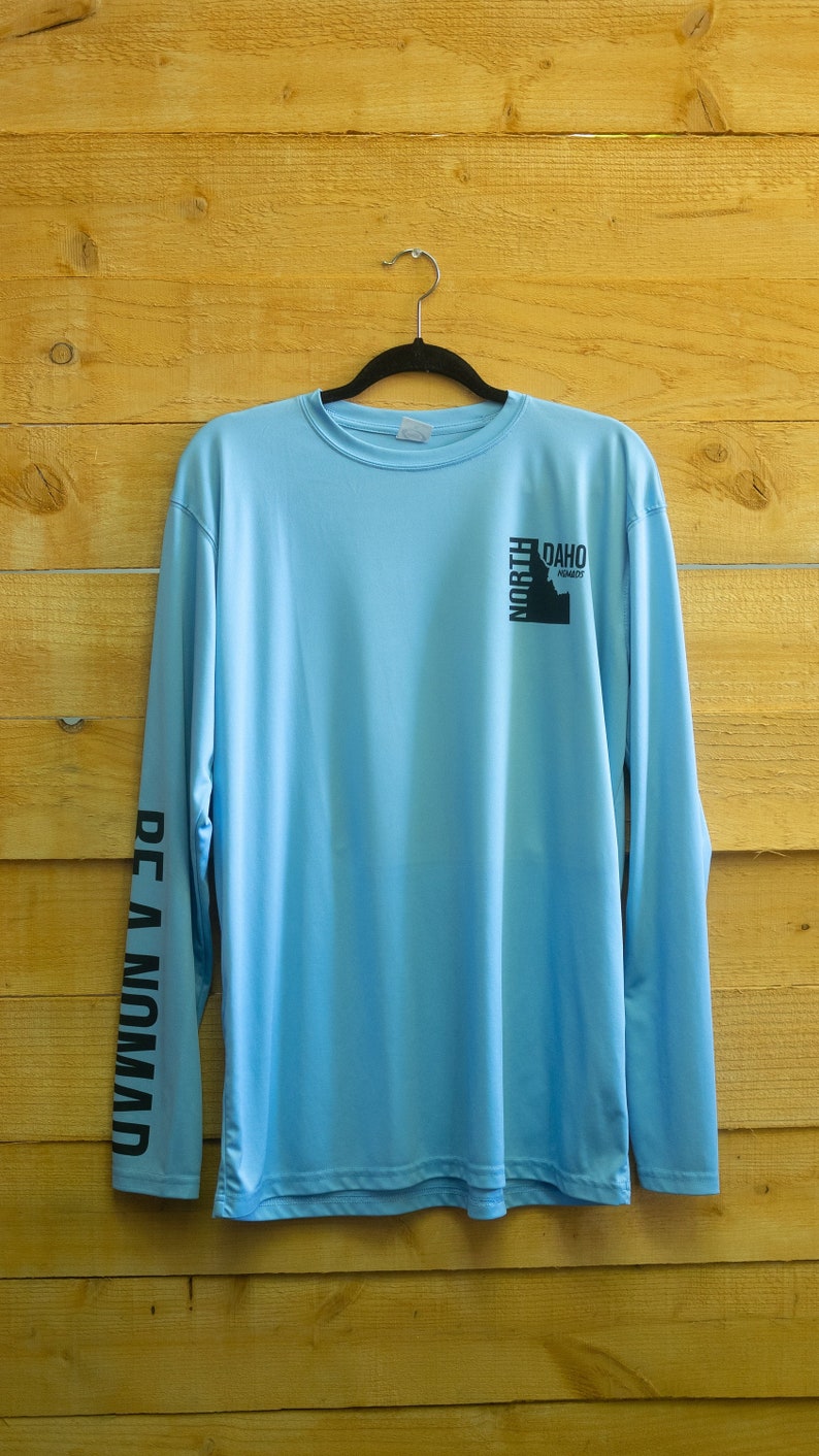May include: A light blue long-sleeved shirt with black text on the sleeves that reads "BE A NOMAD" and a black graphic on the right side that reads "NORTH IDAHO" with a silhouette of the state of Idaho.