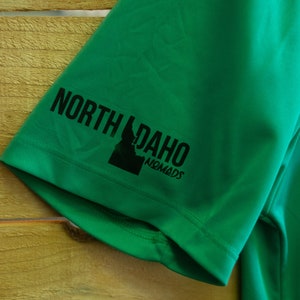 May include: Green short-sleeved shirt with a black graphic of the state of Idaho and the text "North Idaho Nomads" on the sleeve.