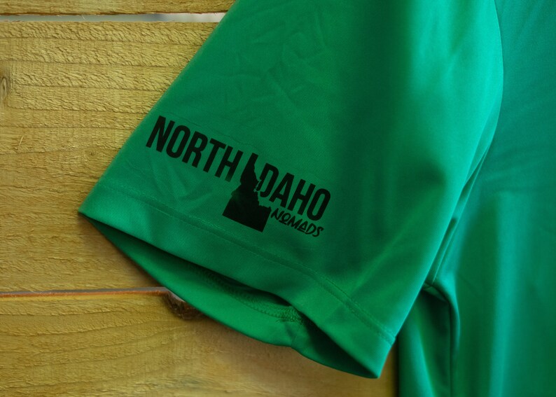 May include: Green short-sleeved shirt with a black graphic of the state of Idaho and the text "North Idaho Nomads" on the sleeve.