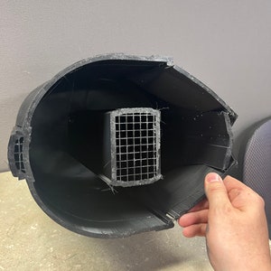 Puede incluir: Un objeto negro impreso en 3D con forma cil&iacute;ndrica curvada. En el interior se ve una estructura rectangular de malla met&aacute;lica. Una secci&oacute;n del objeto est&aacute; abierta, revelando el interior. El objeto est&aacute; sostenido por una mano.