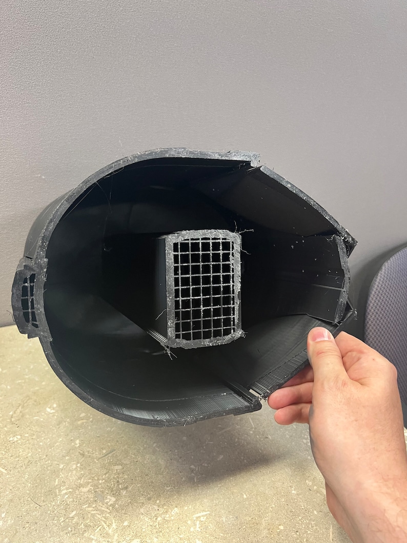 Puede incluir: Un objeto negro impreso en 3D con forma cil&iacute;ndrica curvada. En el interior se ve una estructura rectangular de malla met&aacute;lica. Una secci&oacute;n del objeto est&aacute; abierta, revelando el interior. El objeto est&aacute; sostenido por una mano.